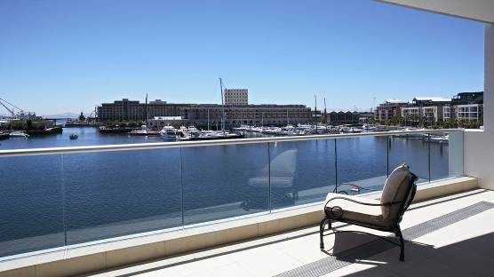 Balcony overlooking harbor with boats and buildings on a sunny day. An empty chair is on the balcony.