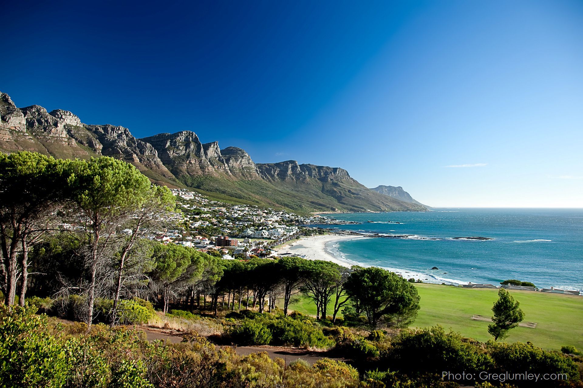 Beach and town nestled at the foot of a mountain range under a clear, blue sky.