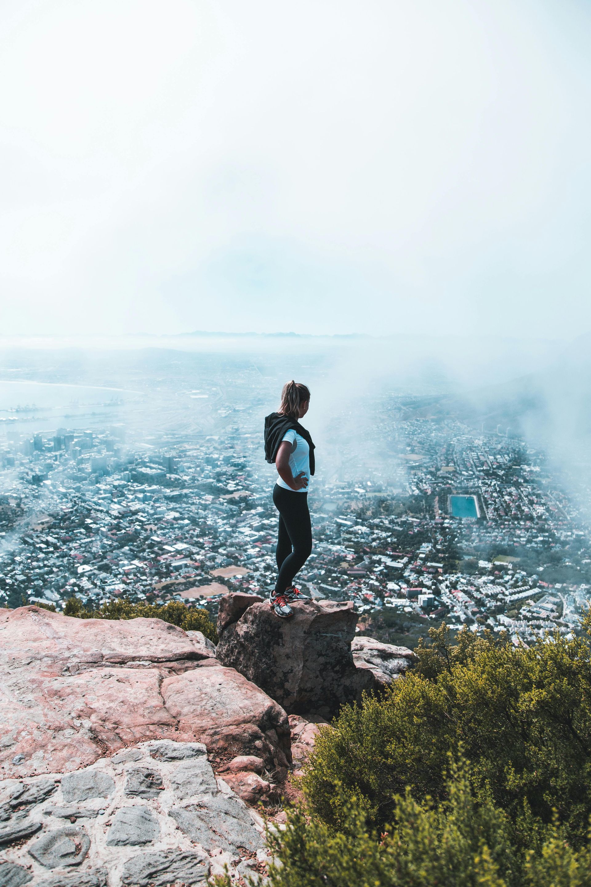 Person on a cliff overlooking a hazy city. They are wearing a backpack and white shirt.