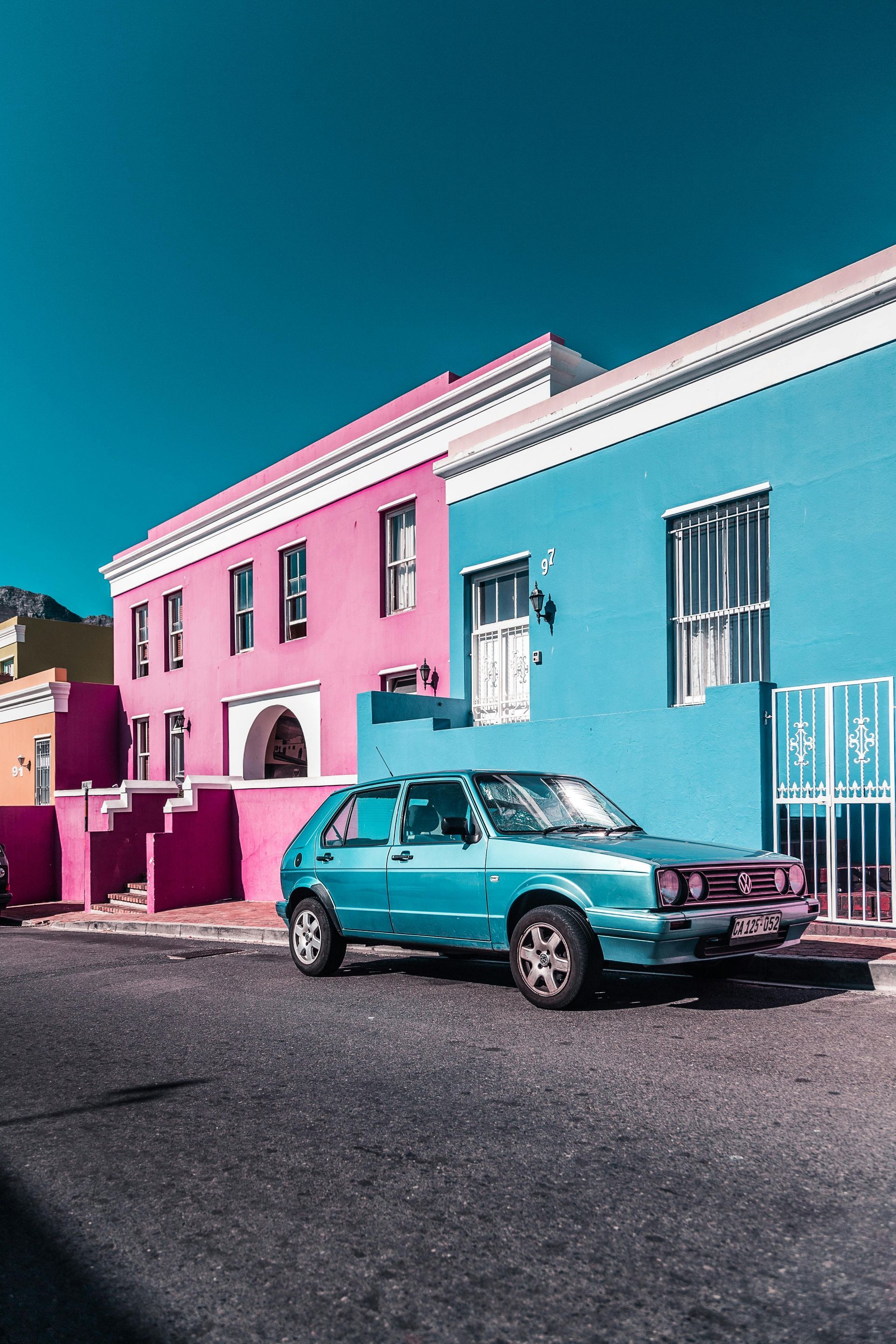 A turquoise car parked on a street in front of colorful houses in pink, blue, and yellow.