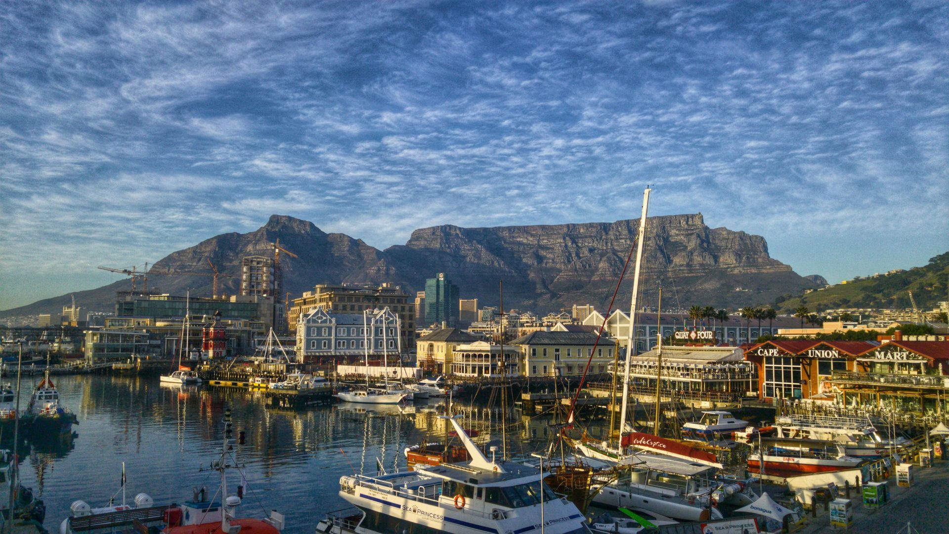 Table Mountain overlooks Cape Town harbor, with boats, buildings, and a cloudy blue sky.