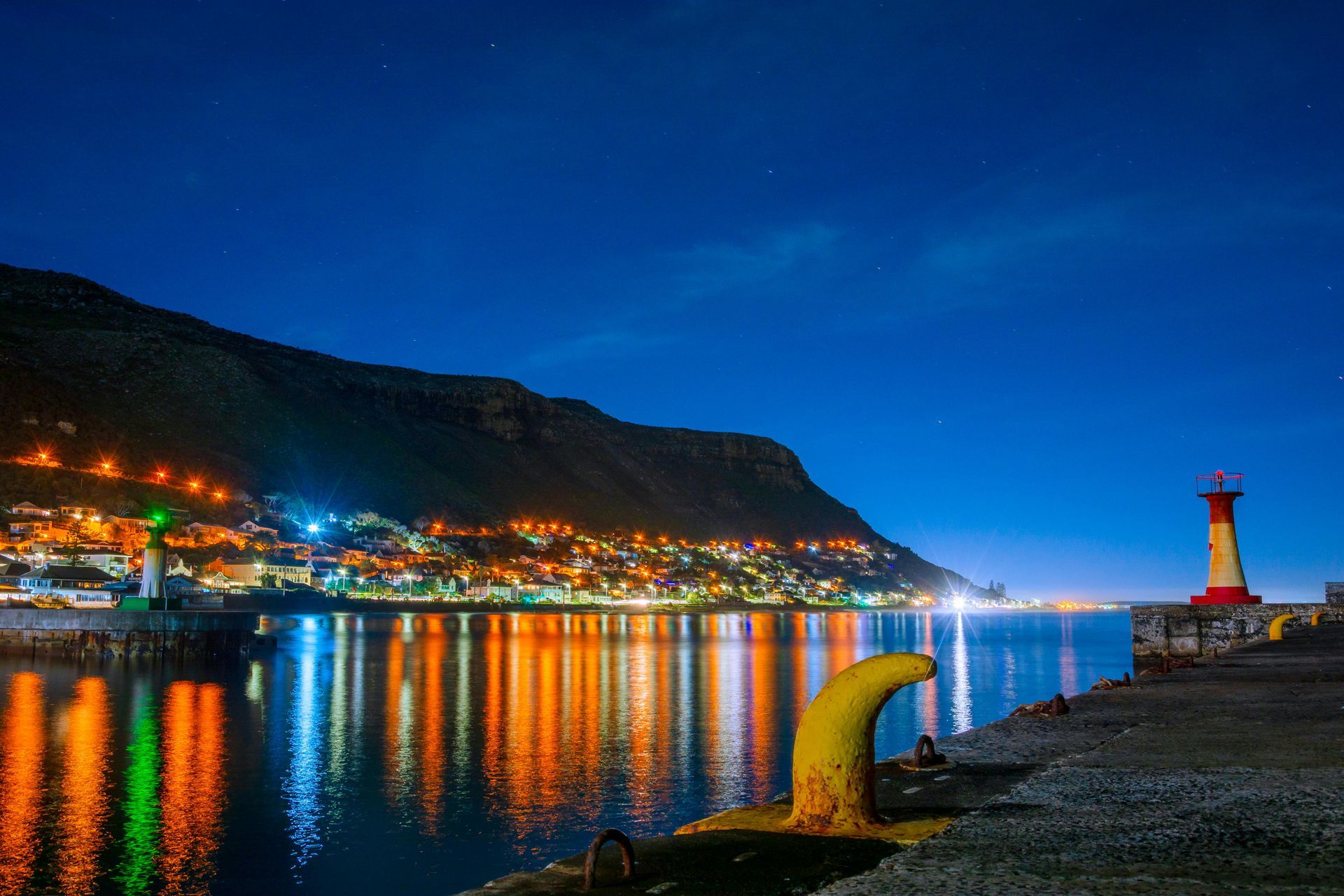 Night view of a harbor town with city lights reflected in the water. A lighthouse stands on the right.