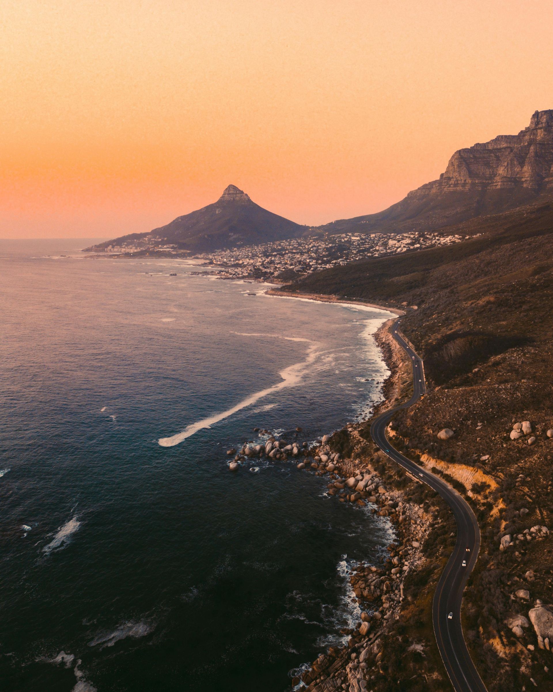 Coastal view at sunset; ocean waves, road, mountains, and orange sky in Cape Town, South Africa.