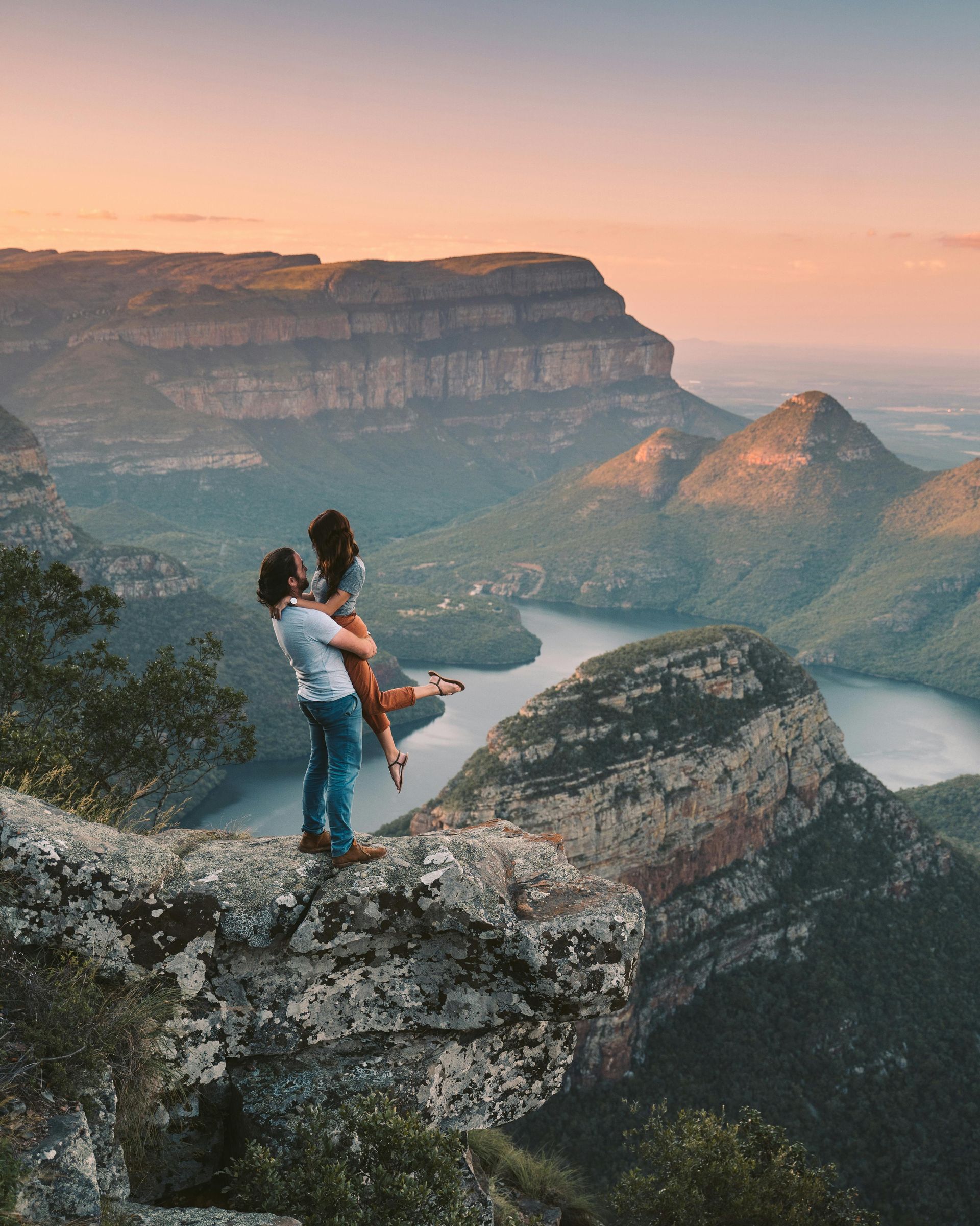 Man lifting woman on a cliff overlooking a river and mountains at sunset.