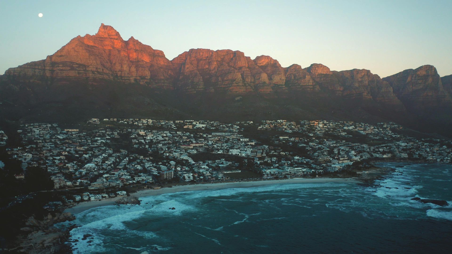 Coastal cityscape at sunset with mountain backdrop.