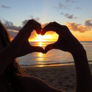 Silhouette hands making a heart shape around a sunset over the ocean.