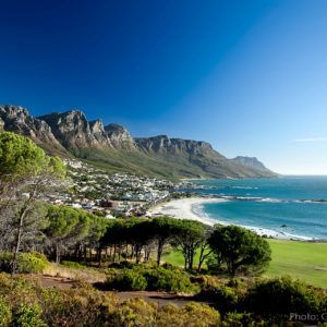 Coastline view of a beach with white sand, ocean, and mountains under a clear, blue sky.