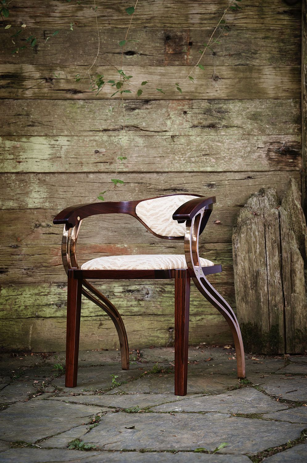 A unique shaped designer wooden chair is sitting in front of a wooden wall.