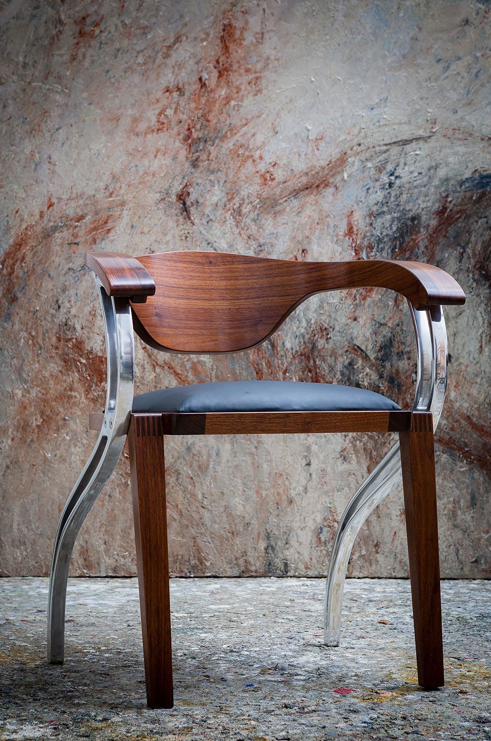 A  unique wooden chair with metal legs is sitting in front of a wall.