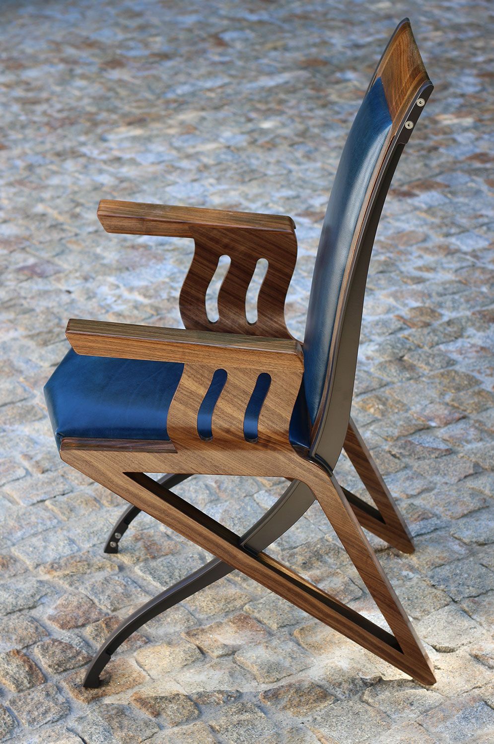 A hand built wooden chair with a blue cushion is sitting on a brick sidewalk.