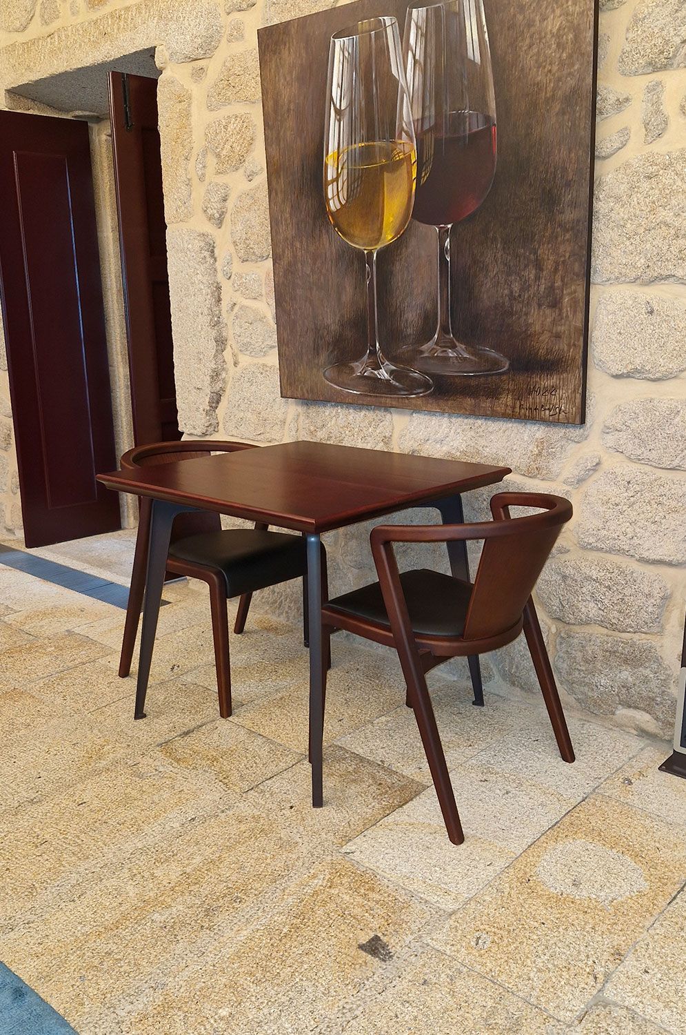 A table and chairs in a room with a painting of wine glasses on the wall.