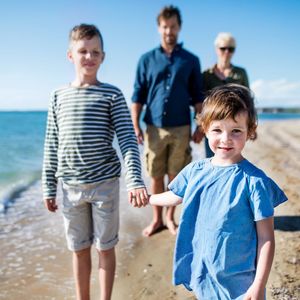 A little girl in a blue dress is holding hands with two boys on the beach.