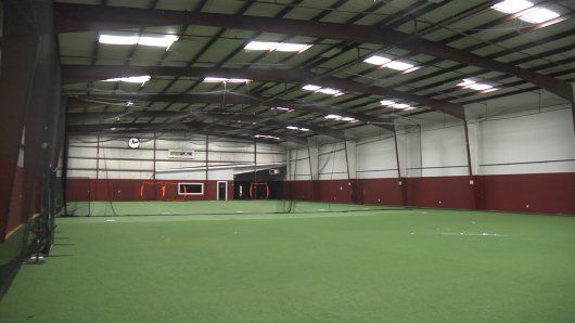 Indoor sports facility with green turf, high ceiling, and burgundy wall base.