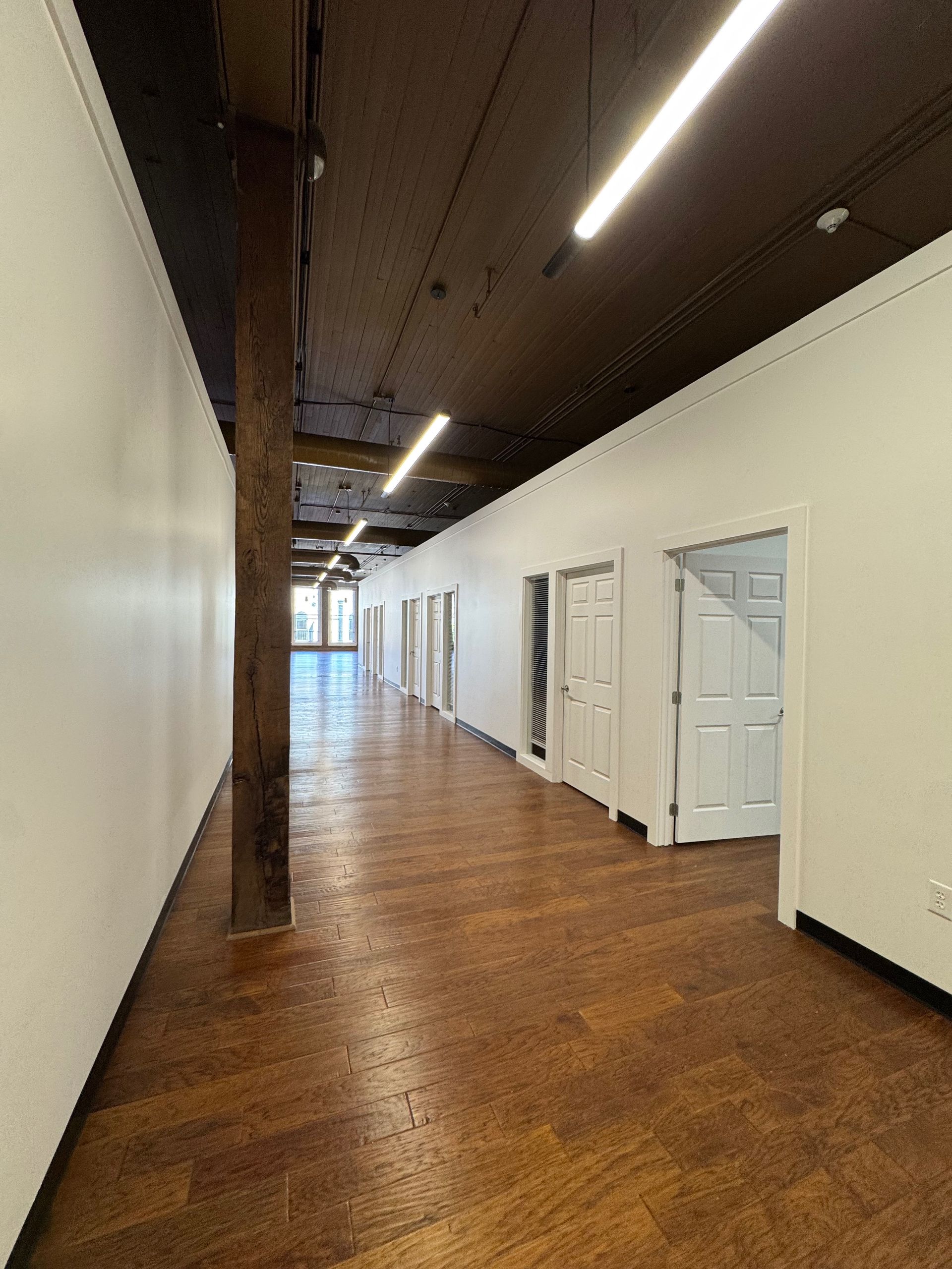 Long hallway with wooden floor and white walls. Doors line the right side, a structural beam on the left.