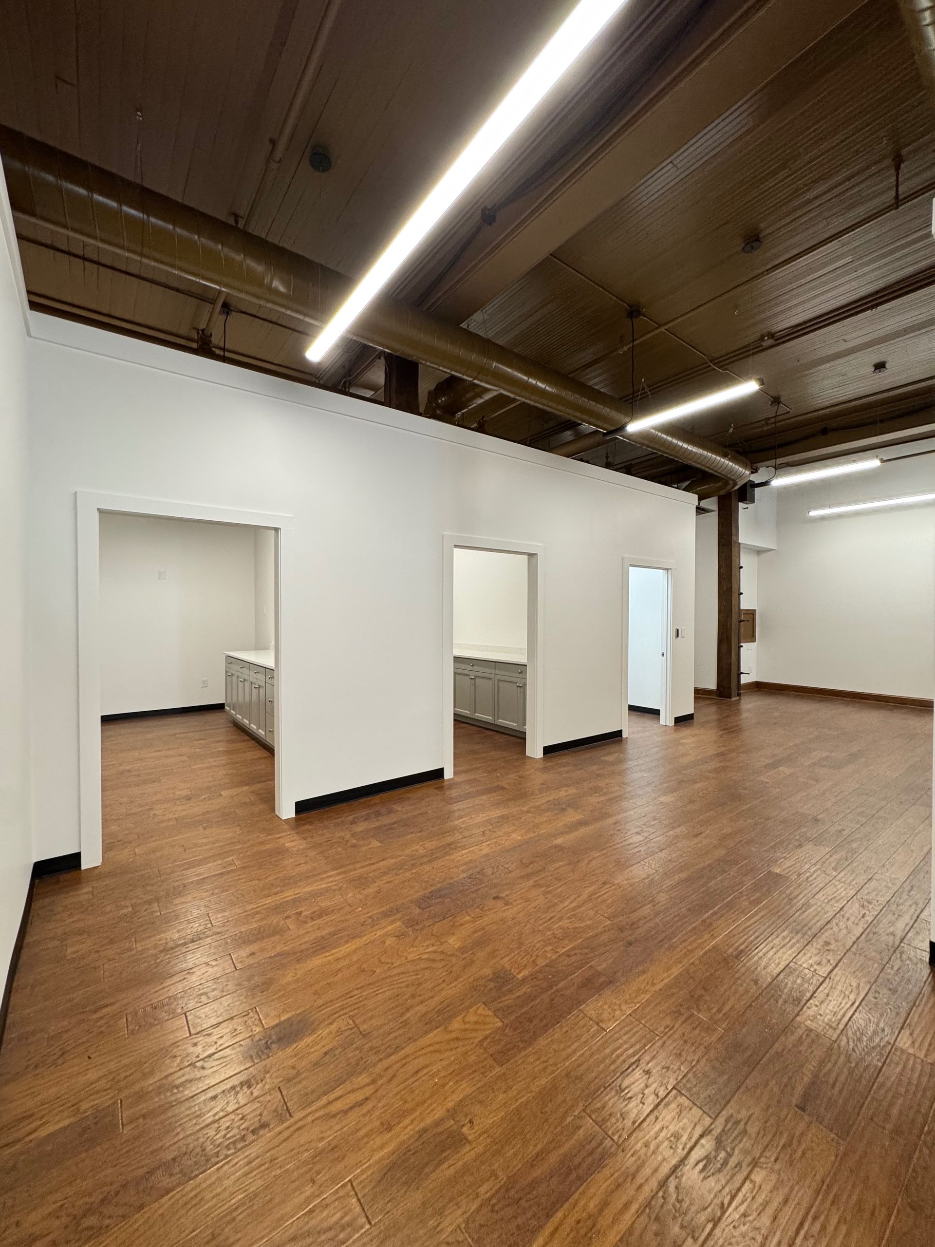 Empty office space with hardwood floors, white walls, and built-in cubicles.