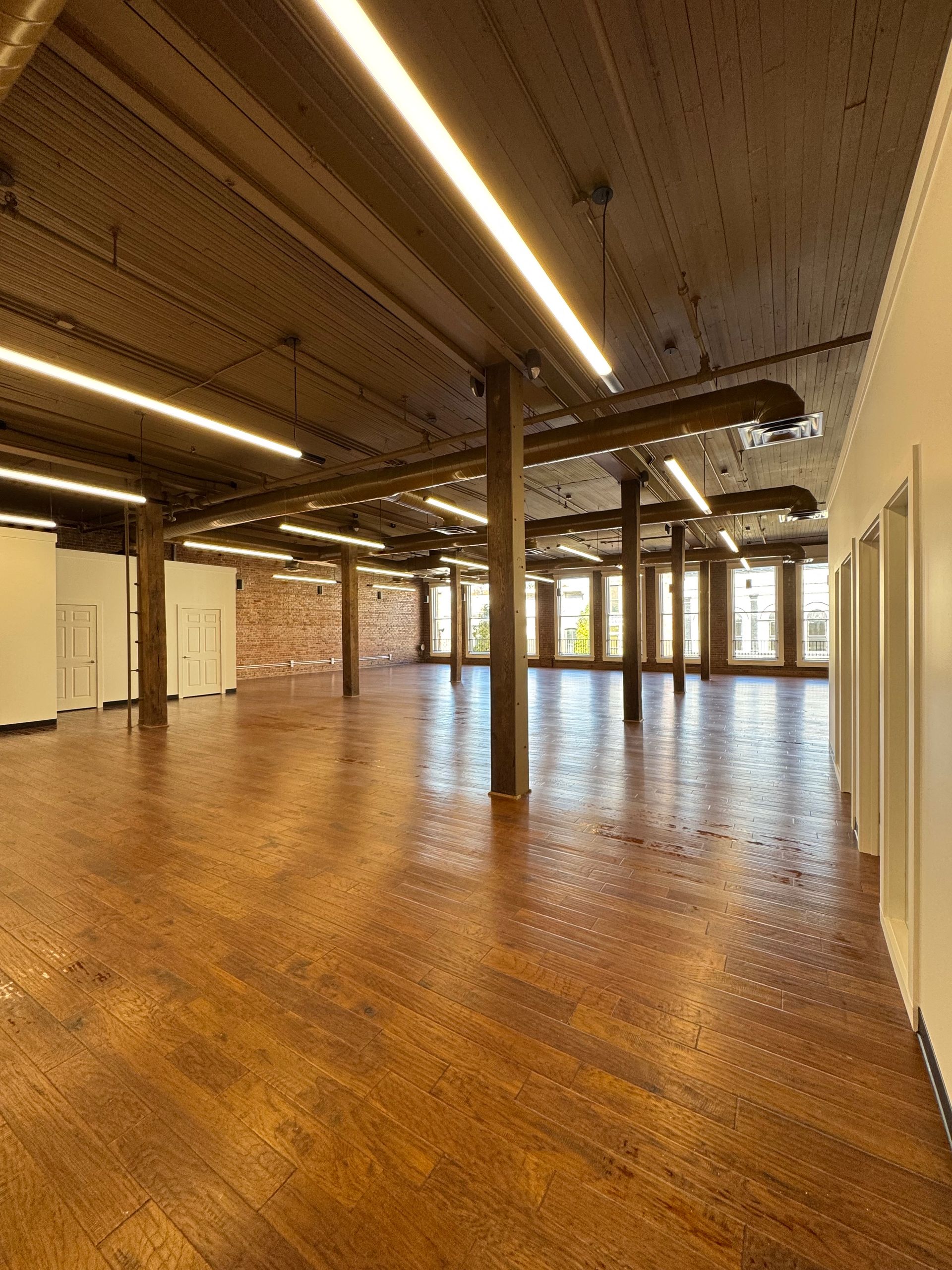 Empty, open-plan space with wooden floor, support beams, and exposed ceiling. Bright overhead lights.