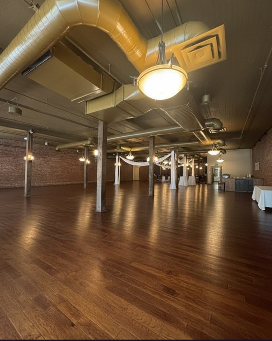 Empty event space with wood floor, brick walls, and decorative ceiling lights.
