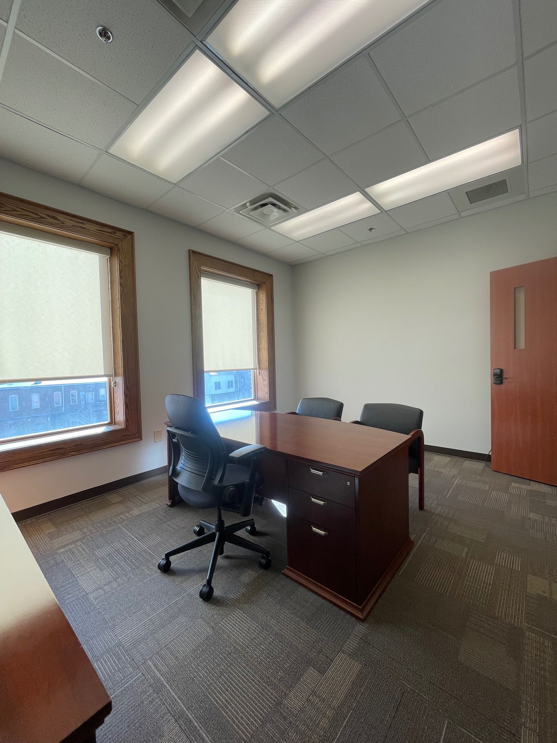 Office with a desk, two windows, two chairs, and a door; grey carpet and white ceiling.