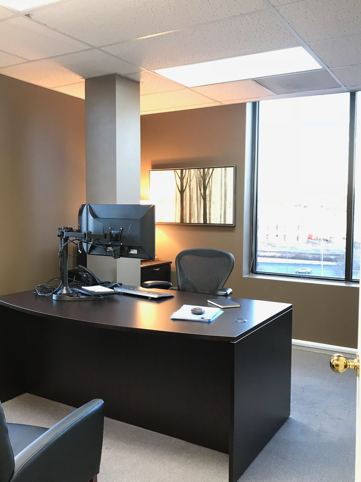 Office with desk, computer, chair, and large window; neutral colors.
