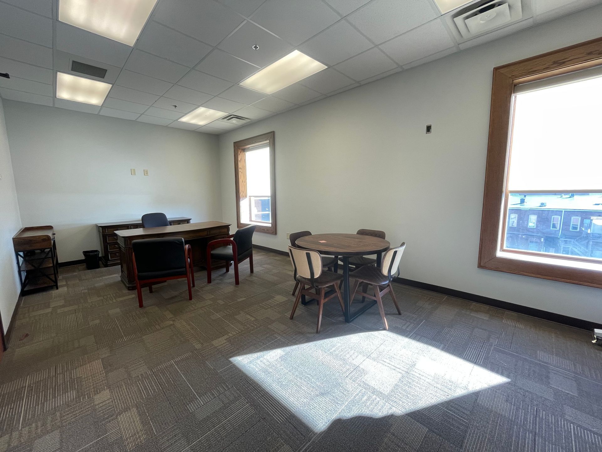 Empty office with desk and chairs, small round table with chairs, carpeted floor, and windows.