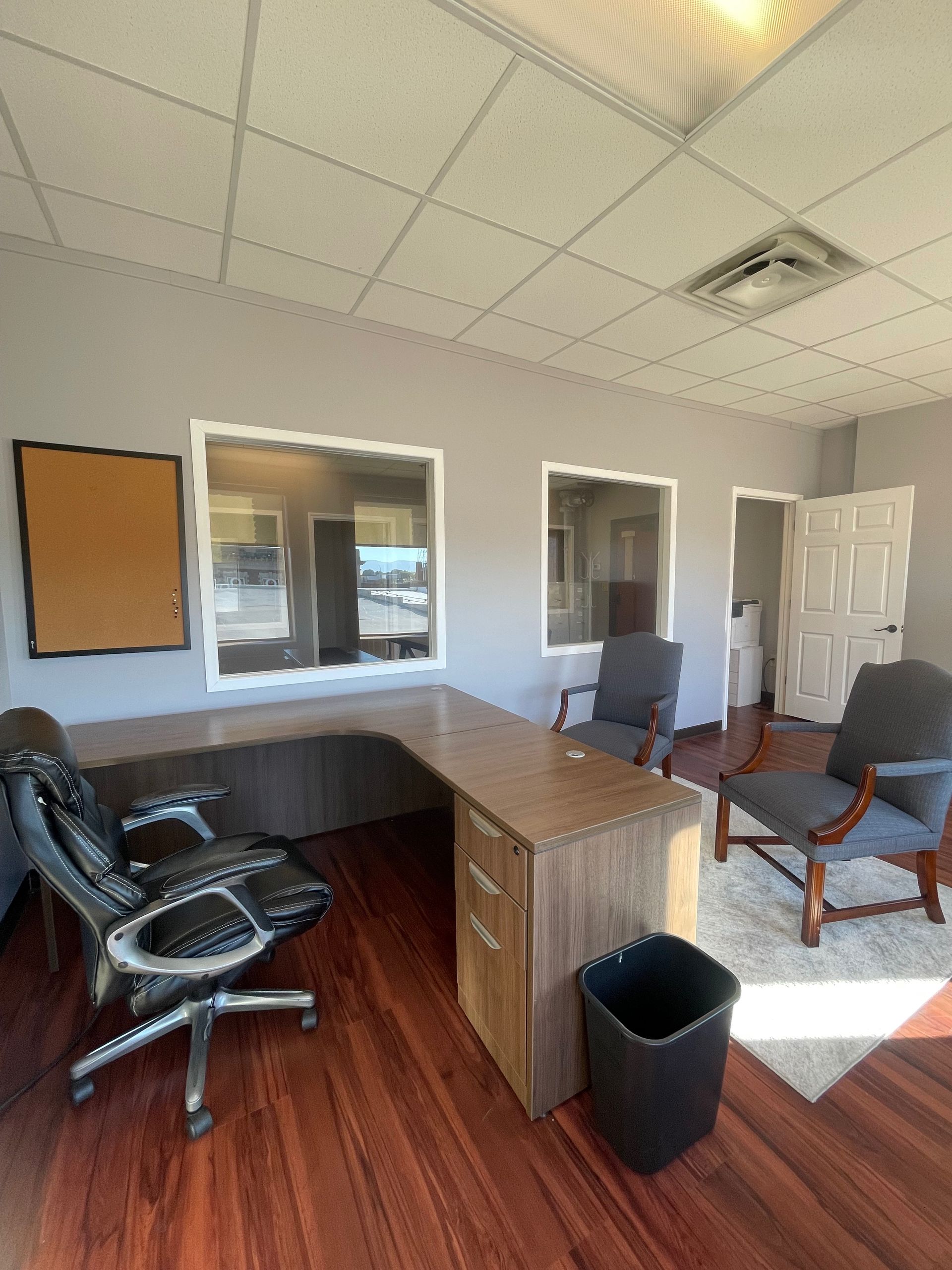 Office with large desk, chairs, and corkboard. Gray walls, wooden floor.