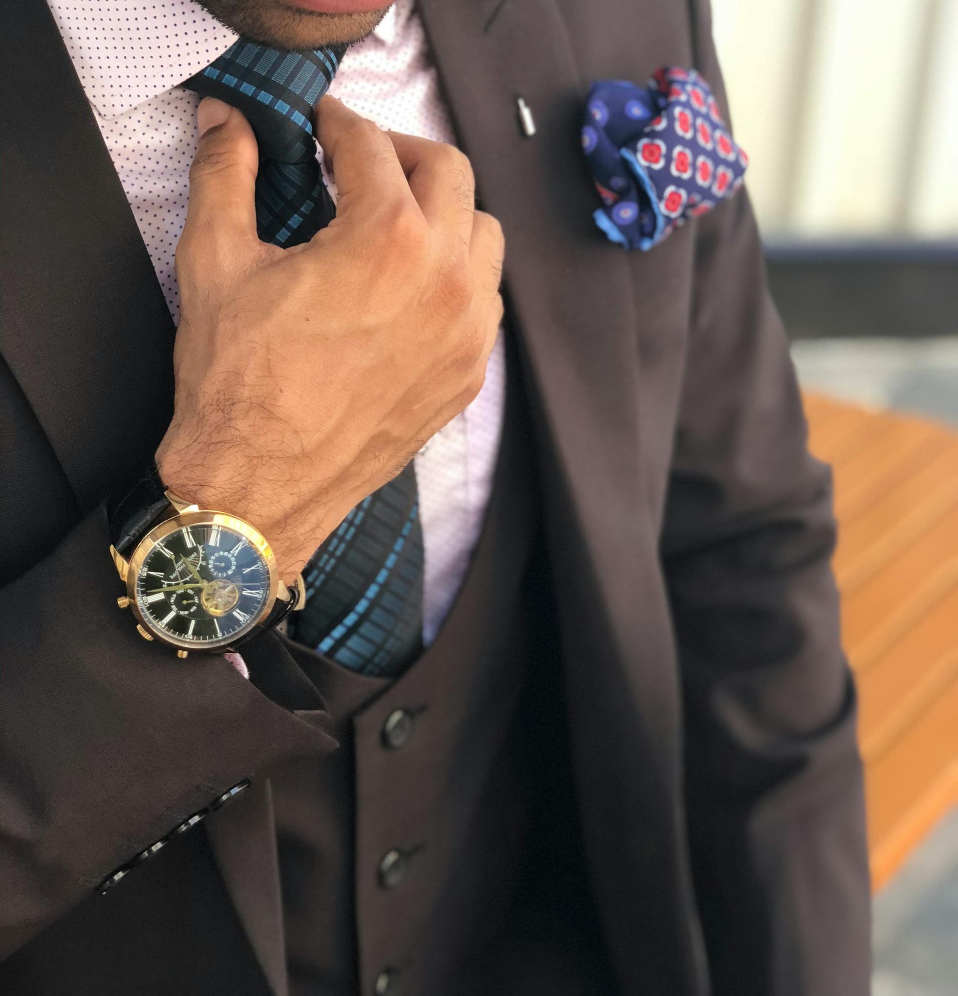 Man in a brown suit adjusting his tie, wearing a watch and a pocket square.