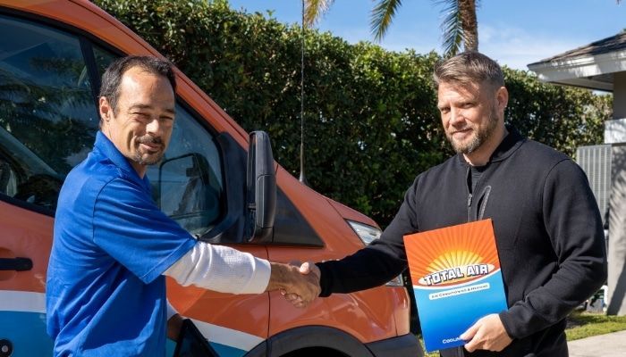 Two men shaking hands in front of an orange van, one holding a brochure.