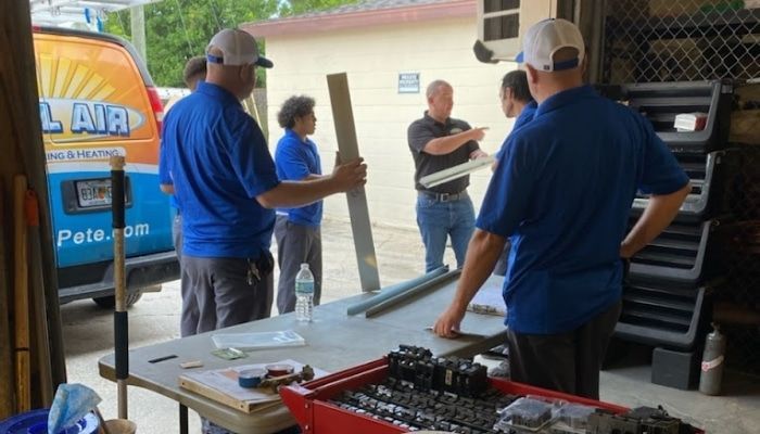 People in blue shirts at a work table, inspecting metal components. A