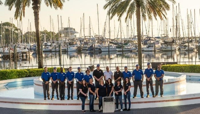 Group of people in blue and black shirts standing near a fountain with boats in the background.