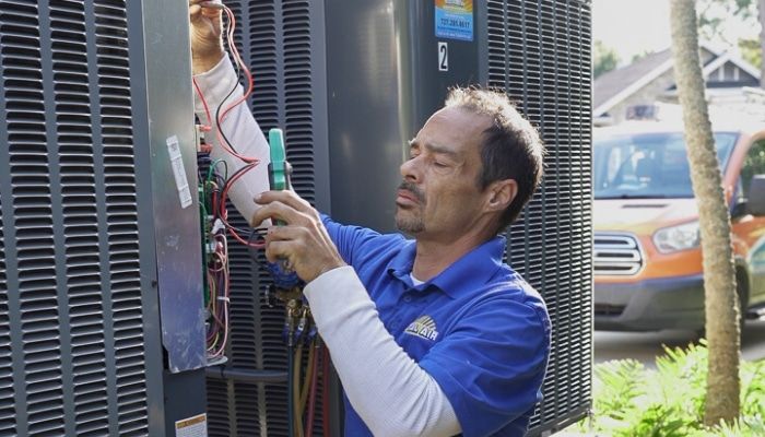HVAC technician in blue shirt wiring an outdoor AC unit; orange service van in background.