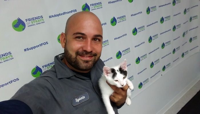 Man holding a small white and black cat, smiling. Green and white logo background.