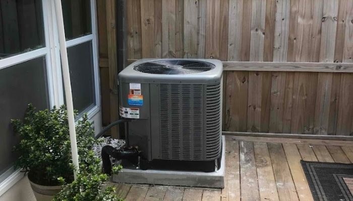 Air conditioning unit outside on a wooden deck, next to a window and a wooden fence.