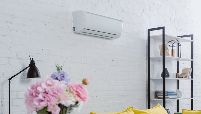 White air conditioner on a white brick wall in a room with a shelf, flowers, and a yellow sofa.