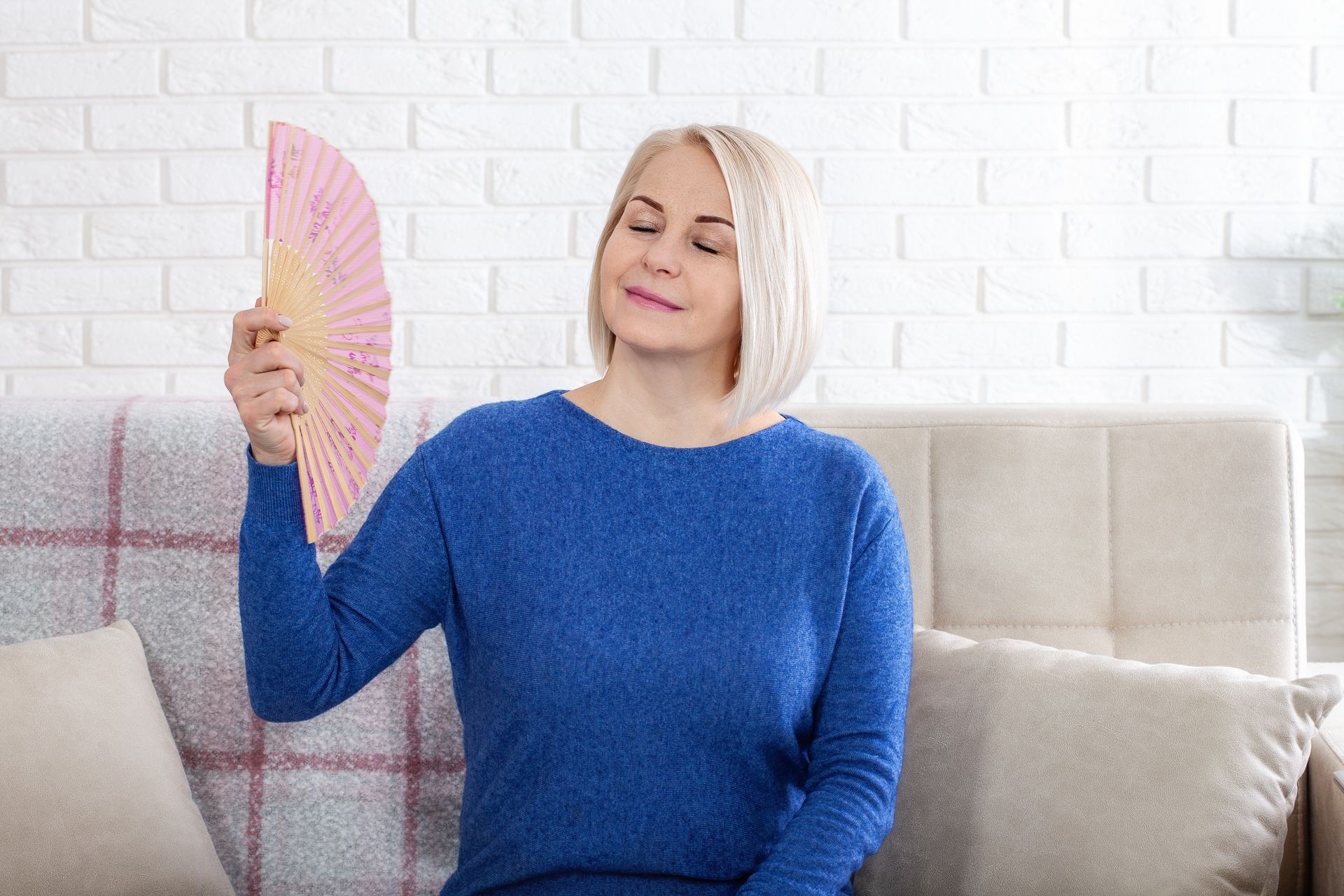 Woman in blue sweater uses pink fan on couch, looking warm.