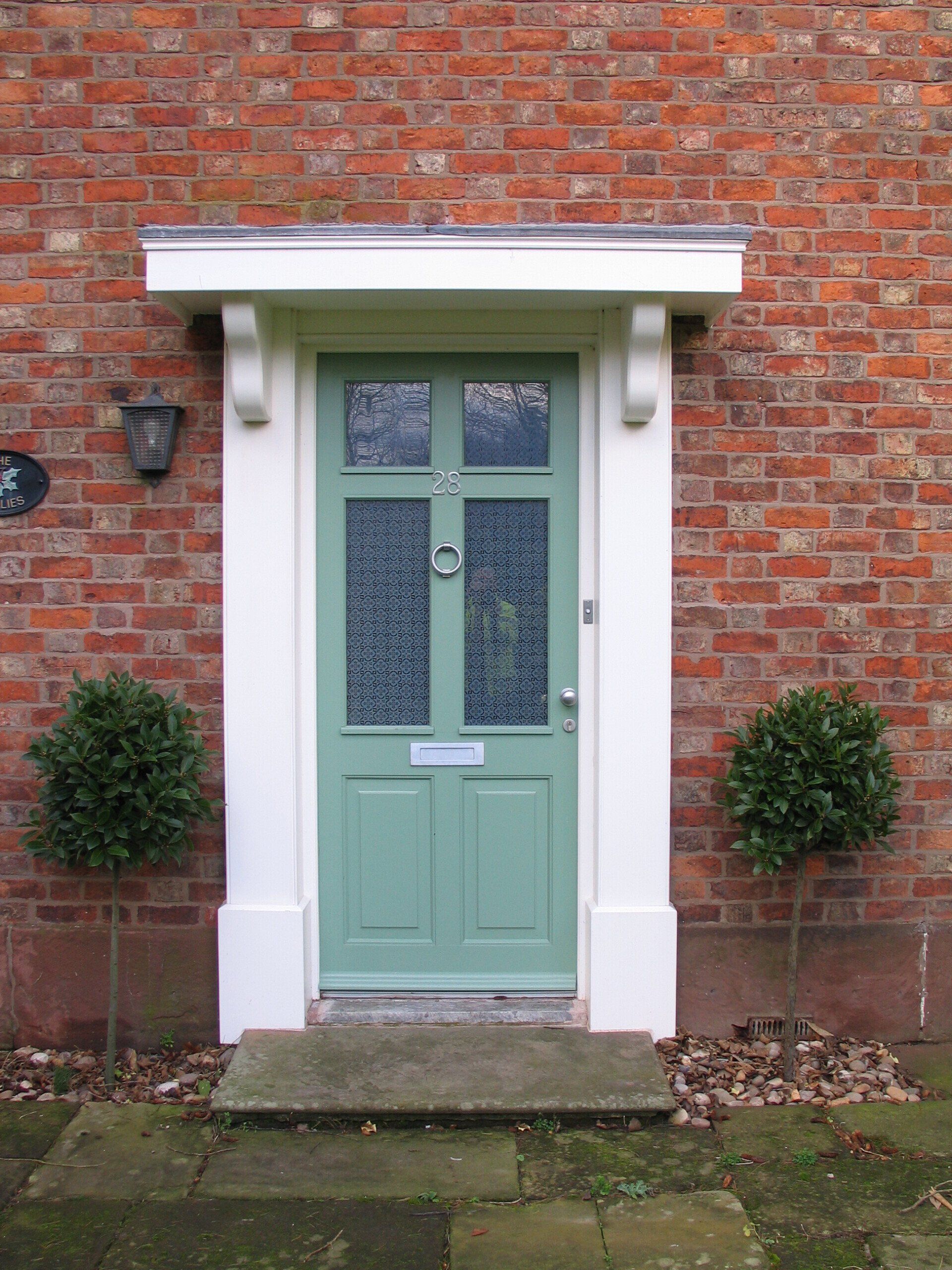 Green door with decorative glass panels and white trim, set in brick wall with two trimmed bushes.