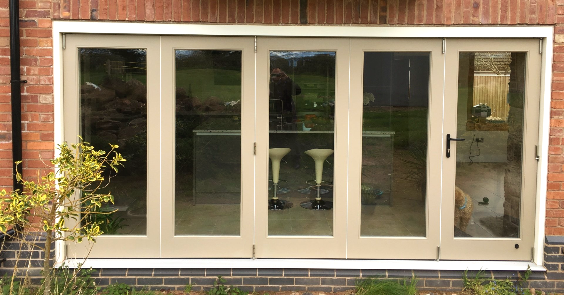 Beige-framed glass bifold doors on a brick house; view of a kitchen with bar stools.