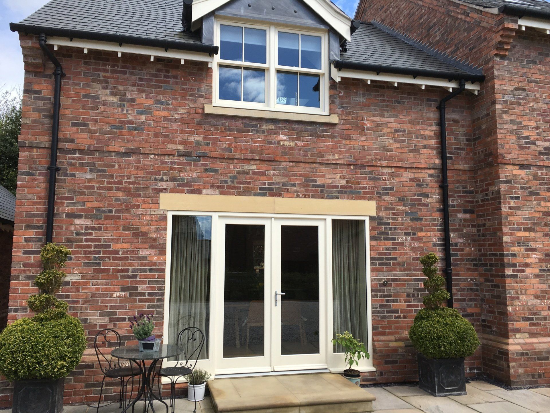 Red brick building with white-framed double doors and window. Black planters with topiary flank the entrance.