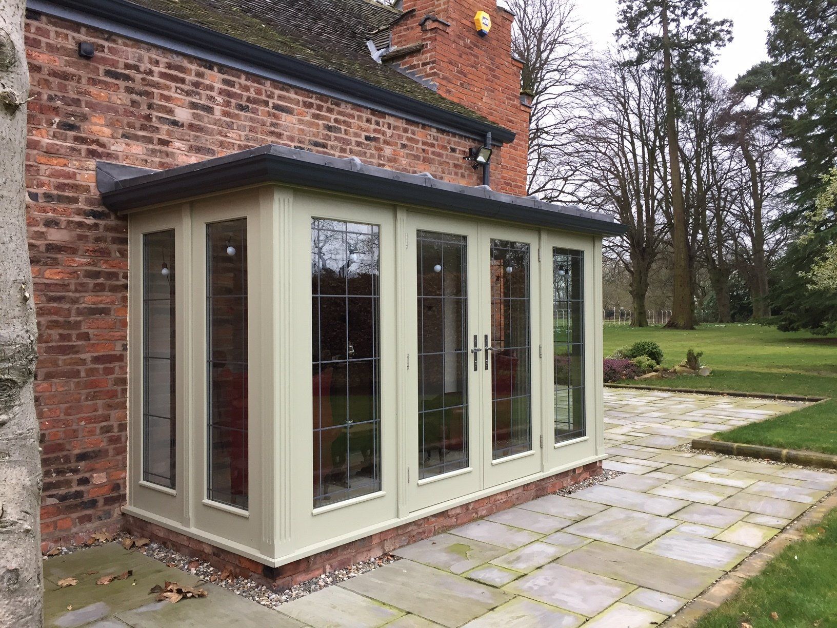 A glass-walled sunroom with green trim attached to a brick building; green grass and a patio in the background.