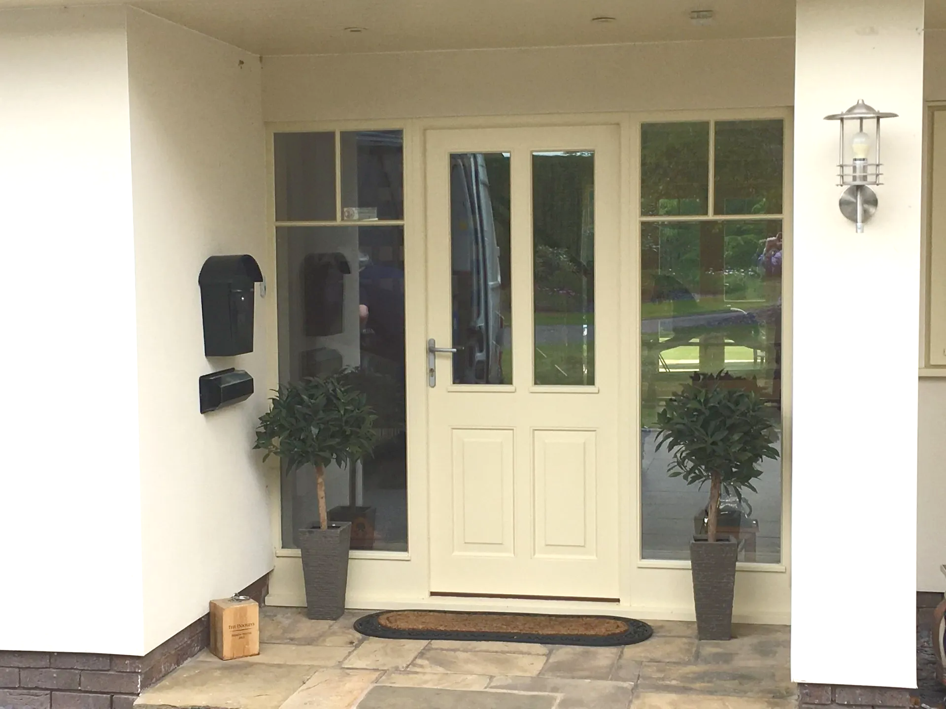 Cream-colored front door with sidelights, mailbox, potted plants, and a welcome mat under a covered porch.