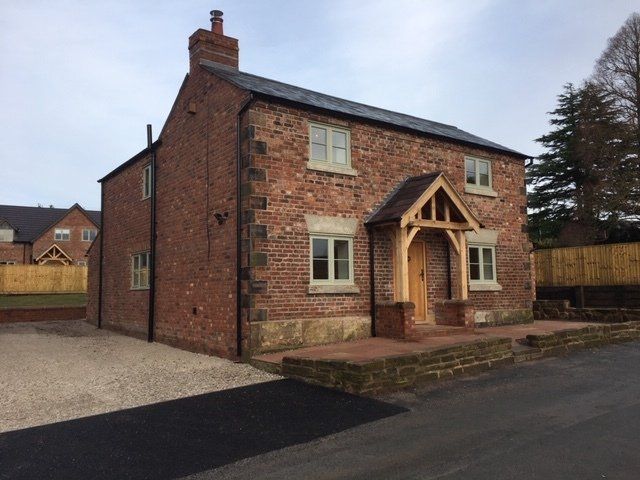 Red brick cottage with oak porch, light green window frames, gravel driveway, and a paved entrance.