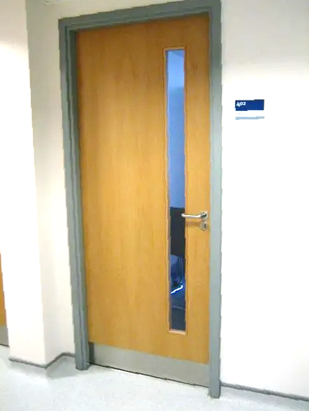 Wooden door with a vertical glass panel, silver frame, handle, and small sign on a white wall.
