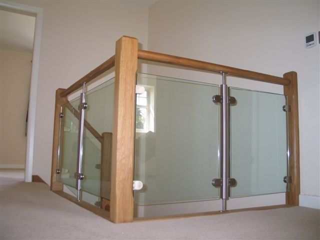 Wooden and glass railing on a landing, with stainless steel supports, light tan carpet, and a window in the background.