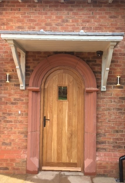Wooden arched door with small window, framed by red stone. White canopy above, flanked by outdoor lights, set in brick.