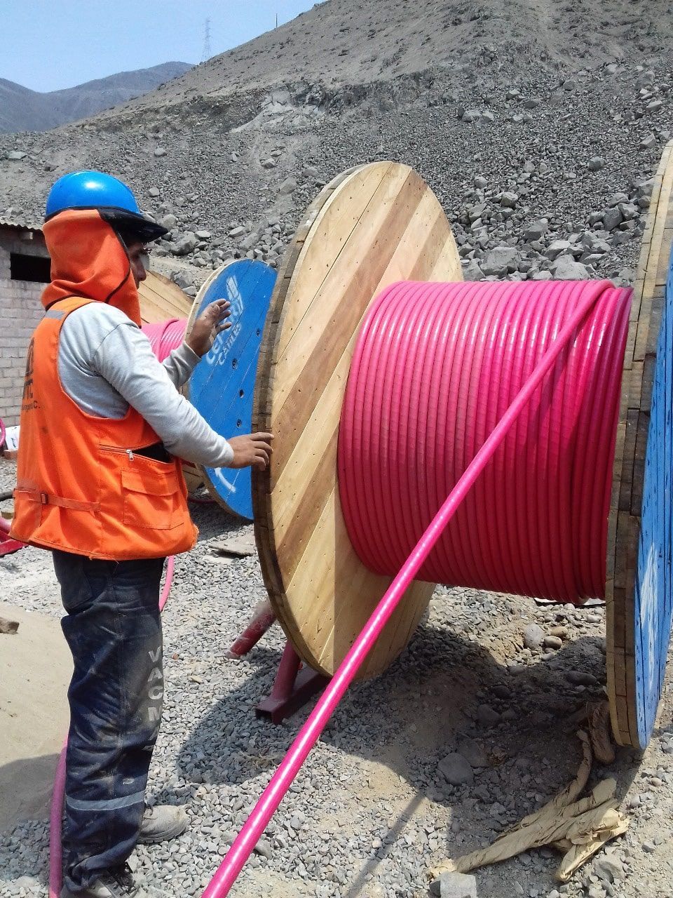 Un hombre con un chaleco naranja está de pie junto a un gran carrete de madera de cable rosa.