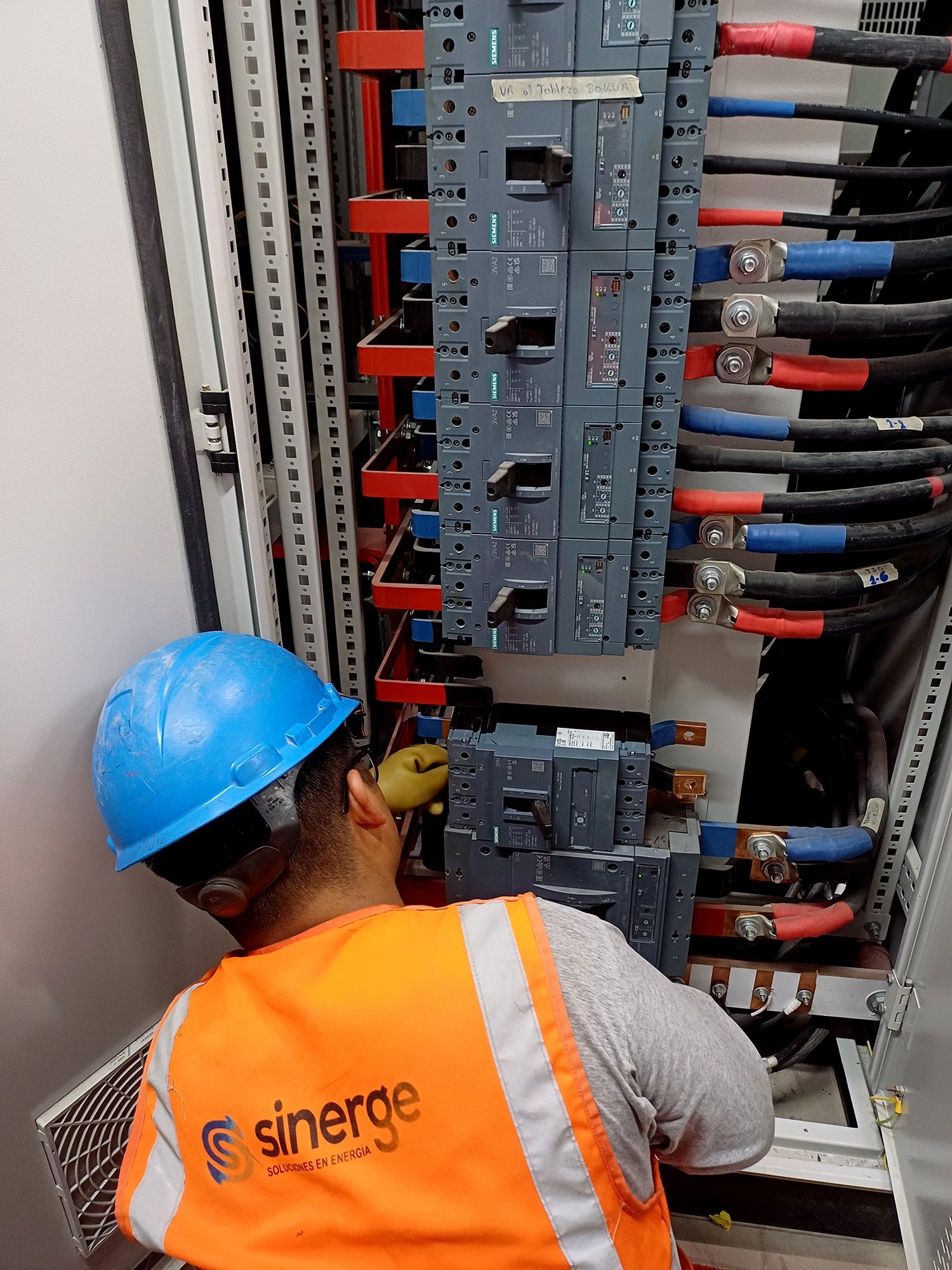 Un hombre que lleva un casco y un chaleco naranja está trabajando en un panel eléctrico.