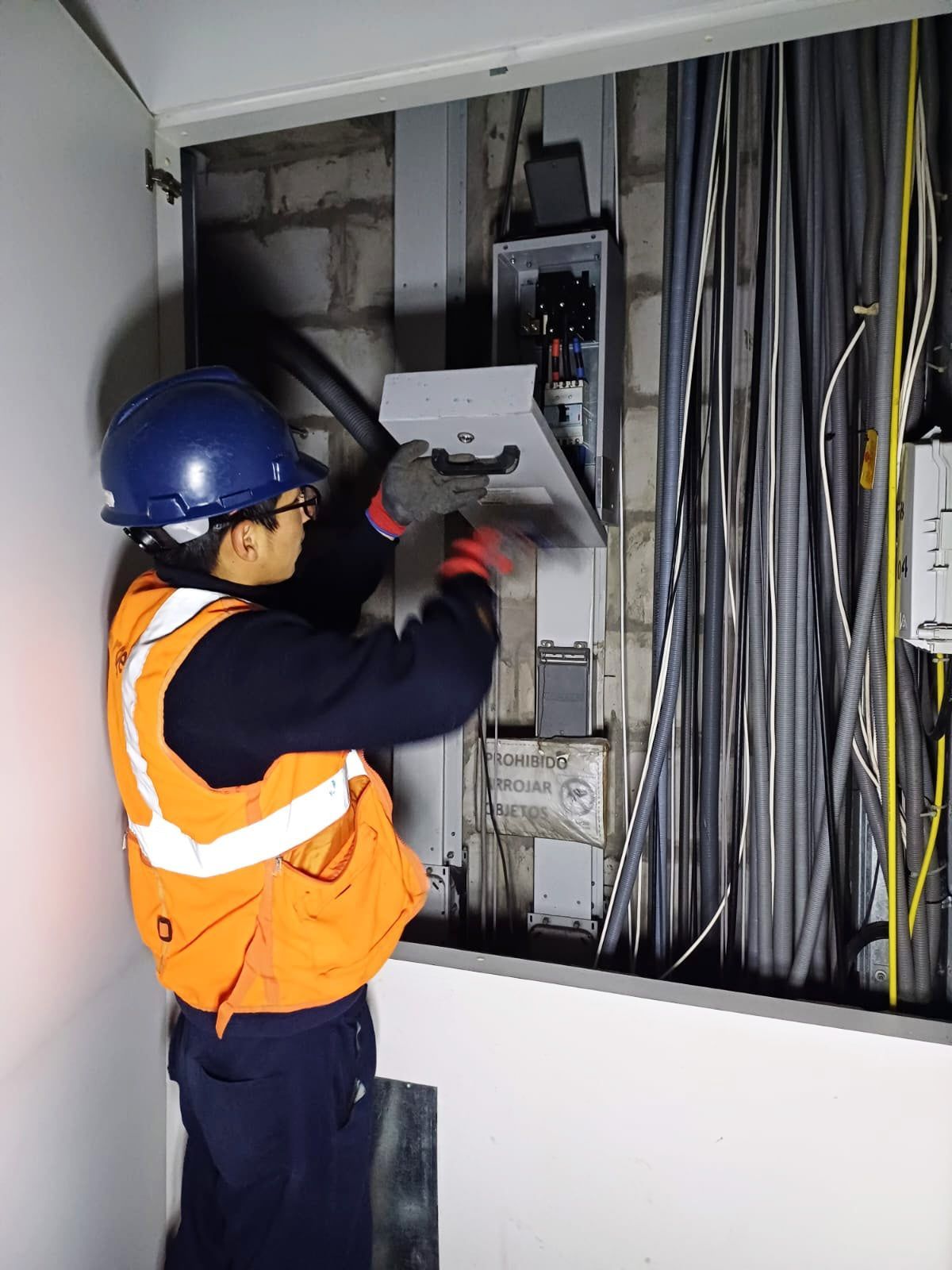 Un hombre que lleva un chaleco naranja y un casco azul está trabajando en una caja eléctrica.
