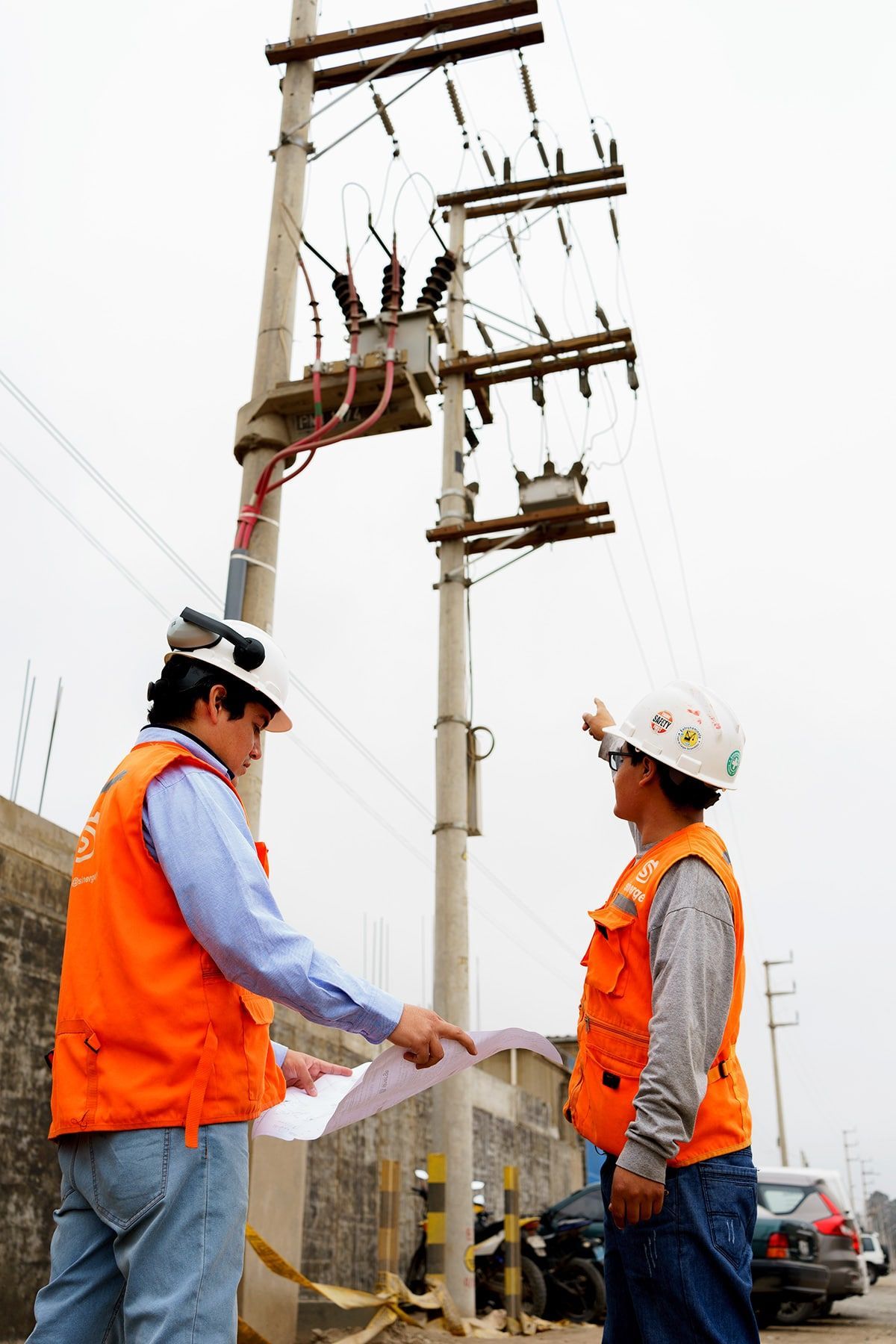 Dos hombres con cascos y chalecos naranjas están de pie junto a un poste eléctrico.