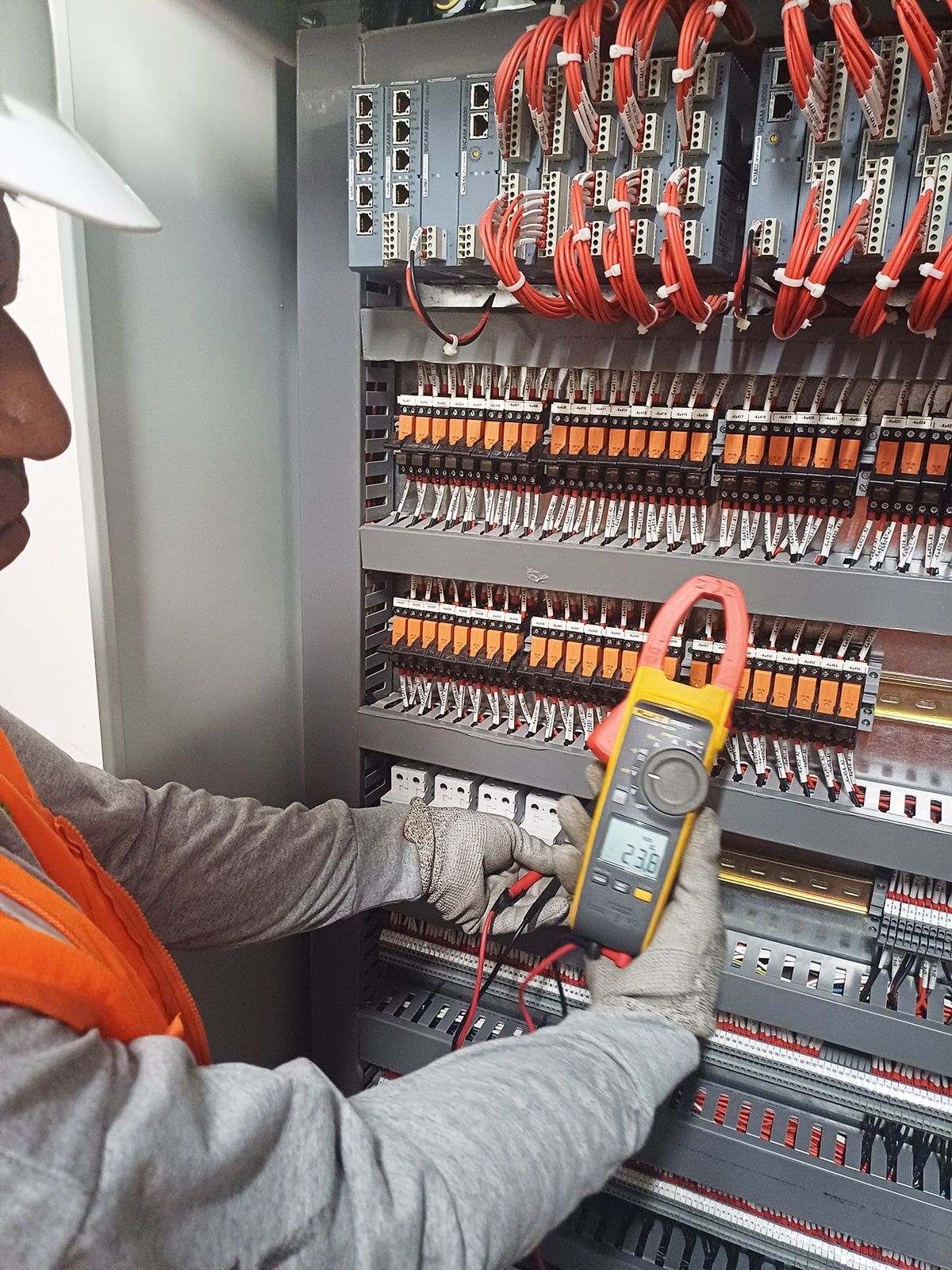 Un hombre sostiene una pinza amperimétrica delante de un panel eléctrico.