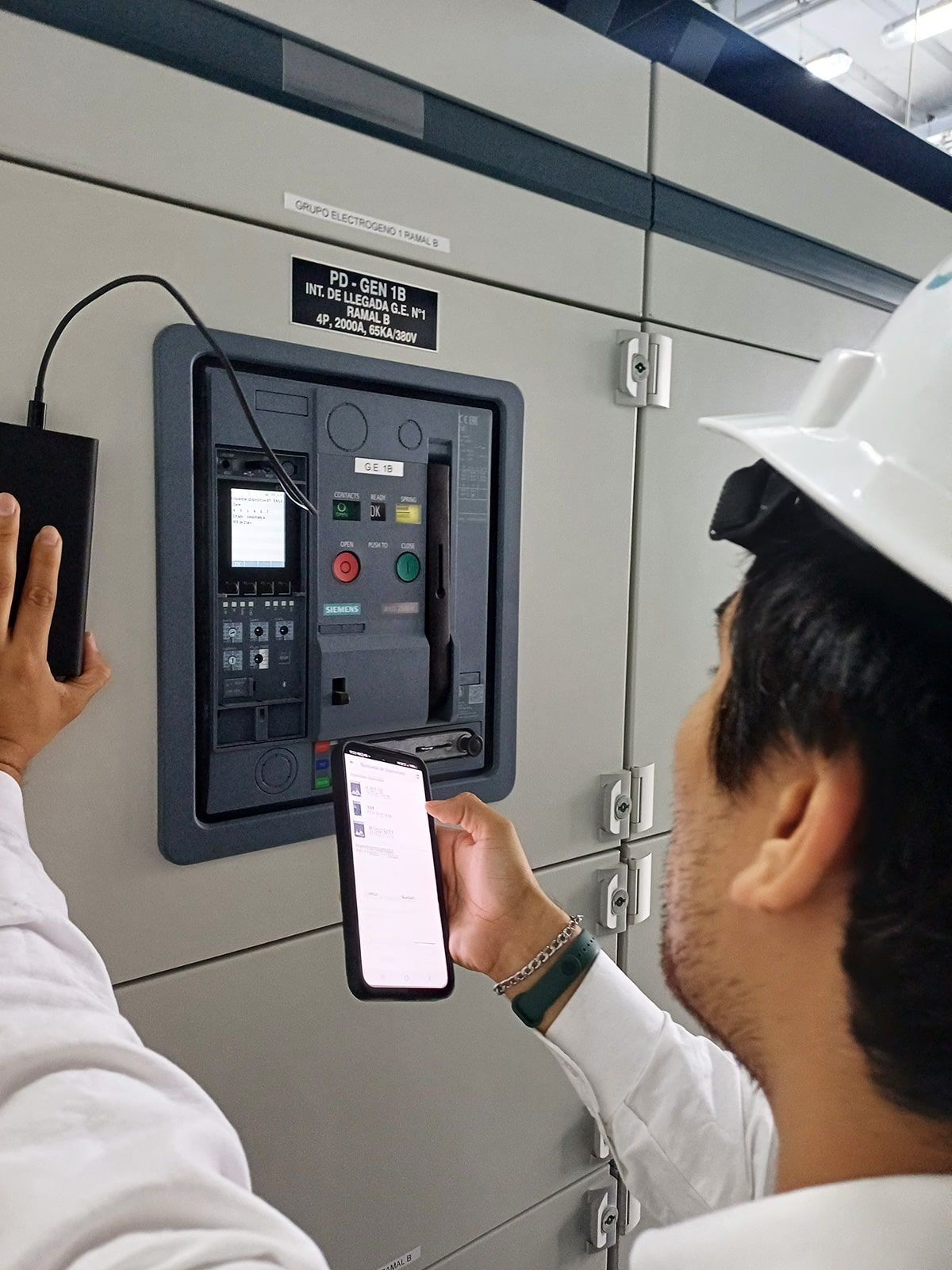 Un hombre que lleva un casco sostiene un teléfono celular frente a un panel de control.