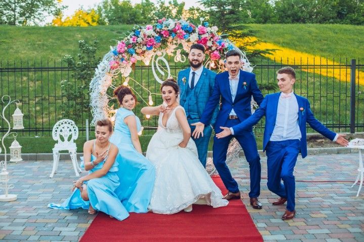 Wedding party posing on a red carpet outdoors with colorful floral decorations and white chairs